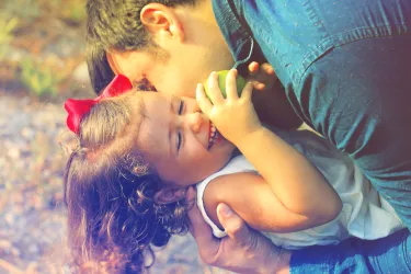 Father lovingly kisses smiling little daughter holding green apple outdoors in warm sunlight.