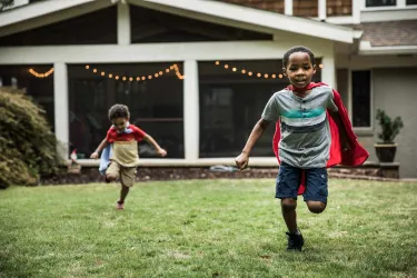 Two young boys wearing red capes running and playing on green grass in a backyard with house in the background