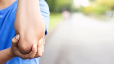 Person in blue shirt holding their elbow, indicating pain or injury, blurred outdoor background.