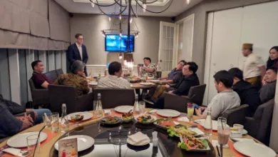 Group of people seated around a large dining table with food, drinks, and a man standing and speaking in a private room