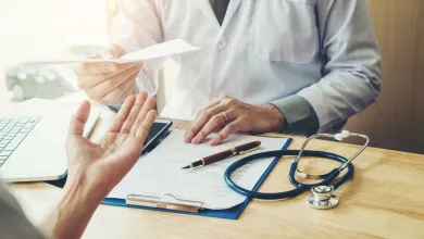 Doctor in white coat handing prescription to patient at desk with stethoscope and clipboard.