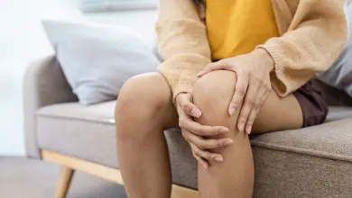 Person sitting on a couch holding their knee in pain, wearing a yellow shirt and beige cardigan.