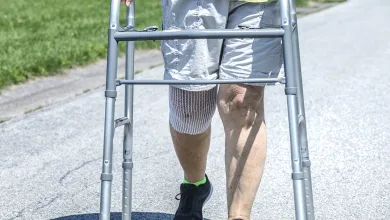 Person using a walker with a knee brace walking on a paved path outdoors in shorts and sneakers.