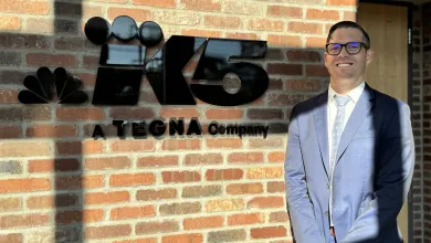 Man in blue suit stands smiling beside black K5 TEGNA Company logo on brick wall in daylight