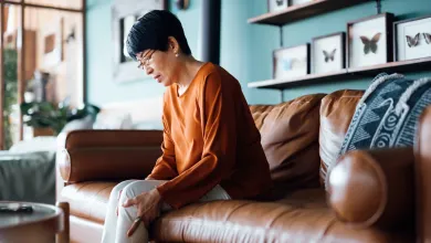 Woman sitting on a brown leather couch indoors, holding her knee, appearing to be in pain or discomfort.