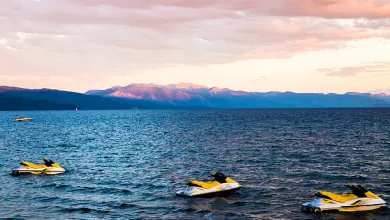 Three yellow and white jet skis floating on a lake with mountain range and pink sky at sunset.