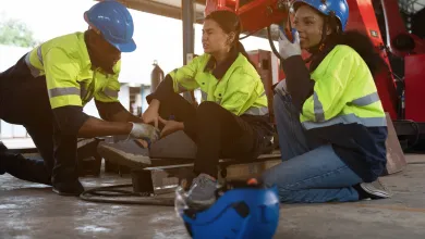 Construction worker attends to injured colleague's ankle while another worker looks on, all wearing safety gear indoors.