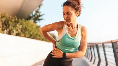 Woman in athletic wear clutching her side in pain while standing on outdoor pathway near water railing.