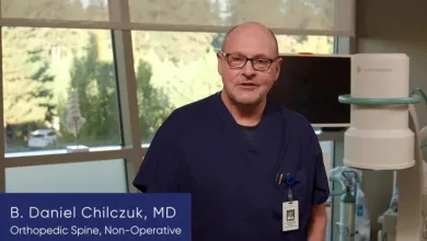 Doctor B. Daniel Chilczuk in navy scrubs standing in a medical office with equipment and trees outside the window.