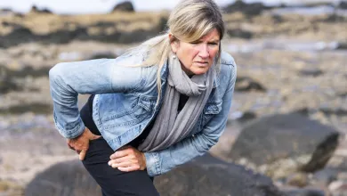 Mature woman outdoors holding hip in pain on rocky shore, wearing denim jacket and gray scarf.