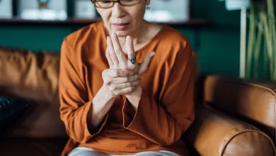 Middle-aged woman in glasses sitting on a sofa, holding and massaging her painful wrist indoors.
