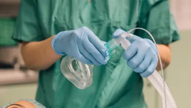 Medical professional in green scrubs wearing gloves adjusts anesthesia mask over patient's face in operating room.