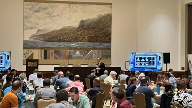 Conference attendees seated at tables in a large room with presenter and screens showing a presentation.