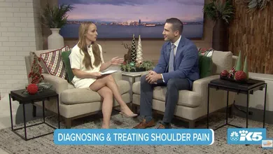 Two hosts discuss diagnosing and treating shoulder pain on a cozy TV set with holiday decor and cityscape backdrop.
