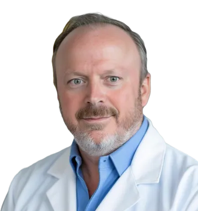 Portrait of a male doctor with beard, wearing a white coat and blue shirt, smiling gently.