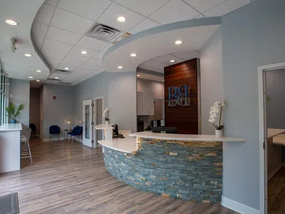 Modern dental office reception area with curved stone desk, wood flooring, blue chairs, and soft lighting.