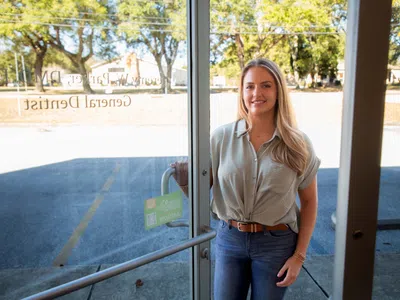 Smiling woman holding door open standing outside a dental office with trees and parking lot in background