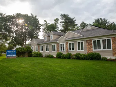 Modern single-story building with stone and siding exterior surrounded by green lawn and trees under cloudy sky.