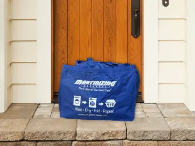 Blue Martinizing branded laundry bag placed on doorstep in front of wooden door and beige siding.