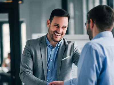 Two men in business attire shaking hands and smiling in a modern office setting during a meeting.