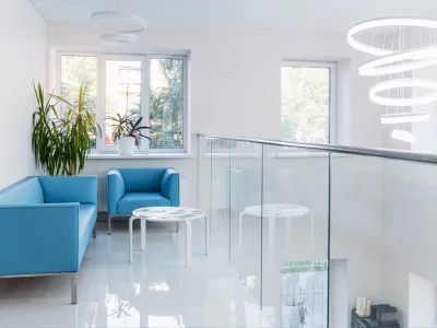 Modern bright waiting area with blue sofas, white round table, large windows, green plants, and spiral LED ceiling light.