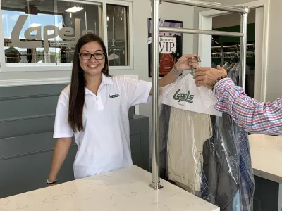 Smiling dry cleaner employee hands over freshly cleaned clothes on a rack to a customer at the counter.