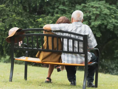 Elderly couple sitting closely together on a park bench with a hat hanging on the bench, surrounded by green trees.