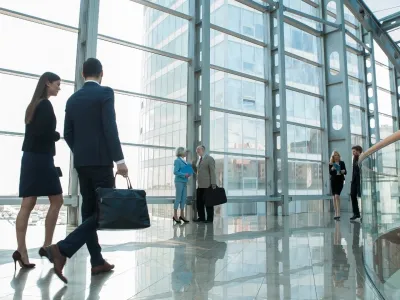 Professional businesspeople walking and conversing in a modern glass office building lobby with natural light.