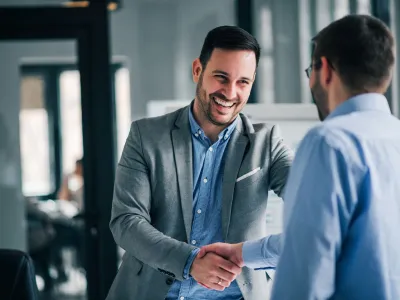 Two businessmen in an office shaking hands and smiling during a professional meeting or agreement.