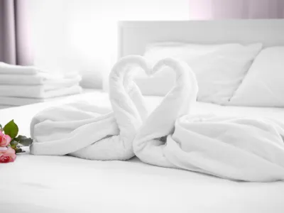 Two white towels folded into swan shapes forming a heart on a bed with pink roses and folded towels in the background
