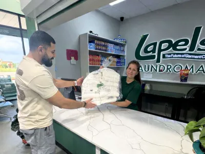 Woman hands a large bundled package of laundry to a man at a laundromat counter with shelving and signage.