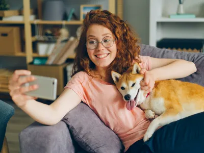 Smiling woman with glasses taking a selfie while holding a happy Shiba Inu dog on the couch at home