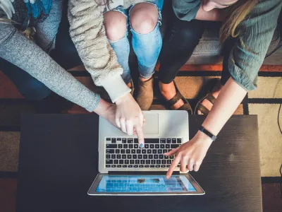 Three people pointing at a laptop screen while sitting on a patterned rug, collaborating closely.