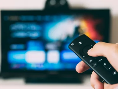 Hand holding a black remote control pointed at a blurred smart TV screen with streaming app icons.