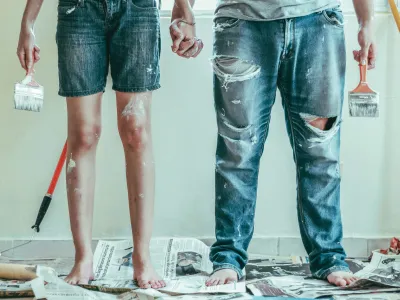 Couple holding hands standing barefoot on newspapers with paint stains, holding paintbrushes in a room being painted