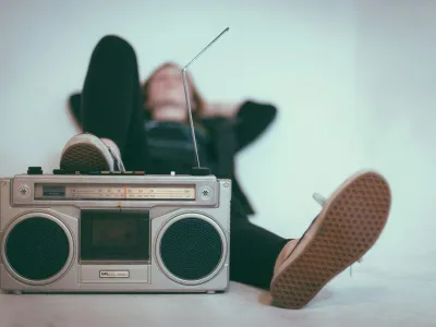 Vintage silver boombox with antenna in focus, person relaxing in background wearing black with sneakers visible.