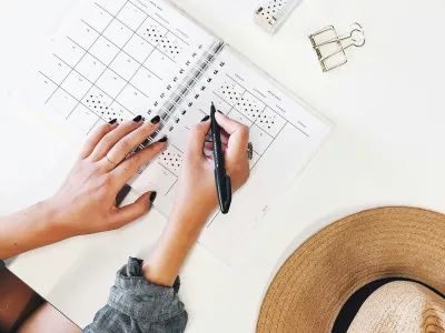 Person writing in a planner with a hat and stationery on a white desk.