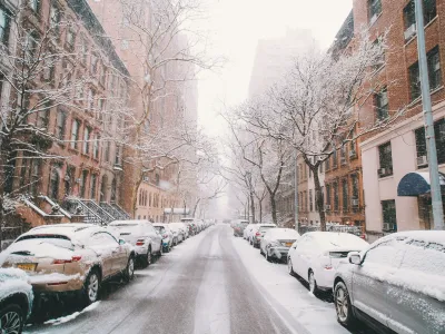 Snow-covered cars and bare trees line a city street with brownstone buildings in winter daylight.