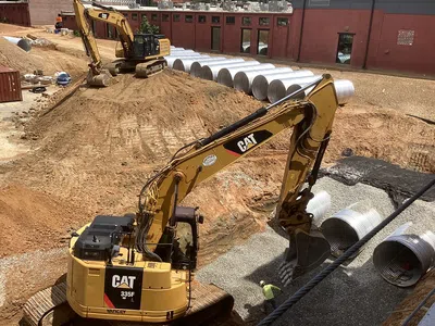 Two CAT excavators working on an urban construction site with large metal drainage pipes and earth moving.