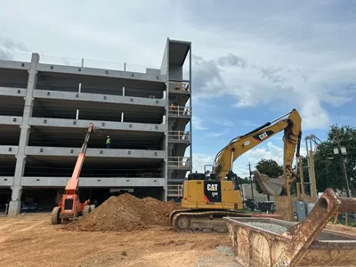 Construction site with a multi-story concrete parking structure, excavator, and workers operating heavy machinery on dirt ground.