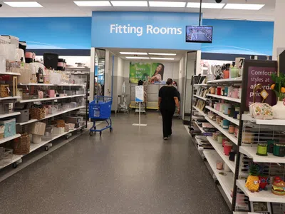 Inside a retail store aisle leading to fitting rooms with shelves of home goods on both sides and a shopper walking ahead.