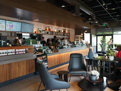Modern coffee shop interior with wooden counters, black leather chairs, customers, and baristas behind the counter.
