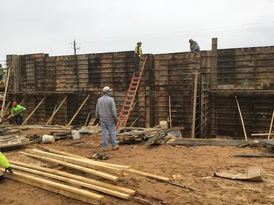Construction workers building a tall wooden concrete formwork wall on a muddy site under overcast sky.
