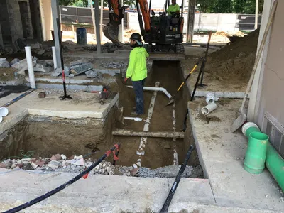 Construction site with workers installing underground PVC plumbing pipes and trench excavation under a building foundation.