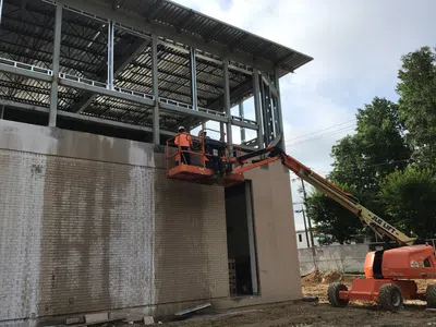 Construction worker on orange aerial lift working on steel frame of building with brick wall and trees in background