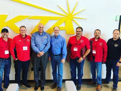 Seven men standing in front of a large yellow KTTA logo on a white wall, dressed in red and blue shirts.