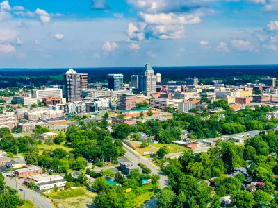 Aerial view of a city skyline with tall buildings surrounded by lush green trees under a partly cloudy sky.