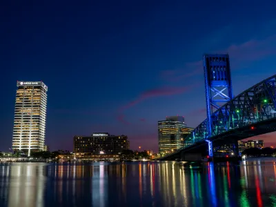 Night view of a city skyline with illuminated buildings and a lit-up bridge reflecting on calm water.