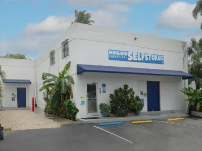 White building with blue awnings and signs for Midgard Express Self Storage surrounded by greenery and parking spaces.