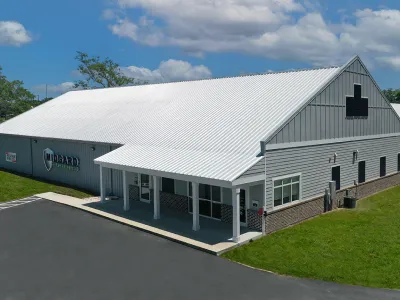 Gray and white commercial building with large metal roof and green lawn under blue sky with clouds.
